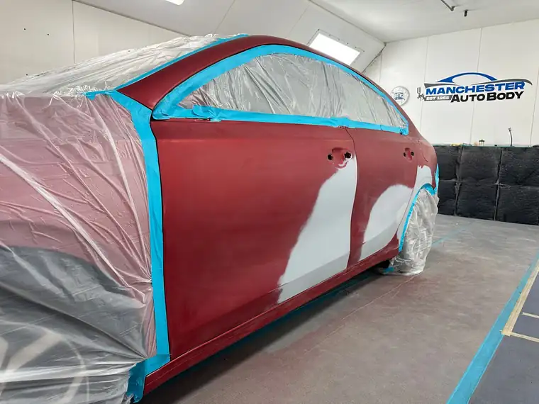 A red sedan masked in the paint booth with primer on the rear door and quarter panel