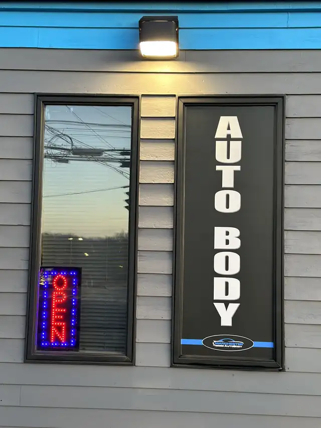 The black vertical AUTO BODY sign and lit OPEN sign on the grey clapboard of the Vernon shop