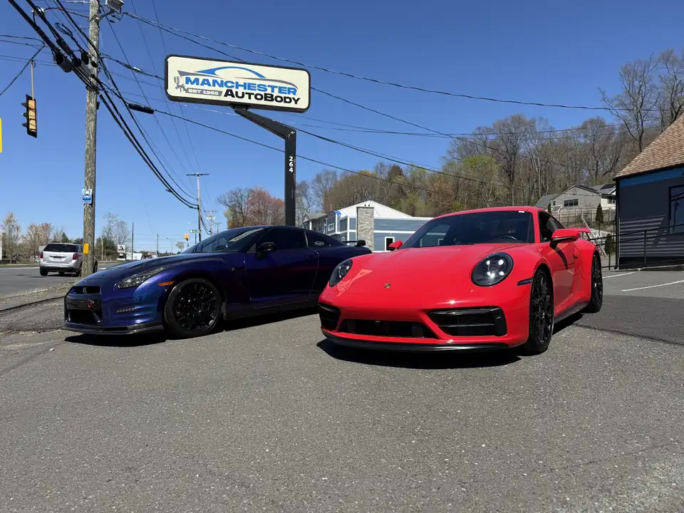 A blue Nissan GT-R and a red Porsche 911 on the lot beside the Manchester Auto Body pole sign
