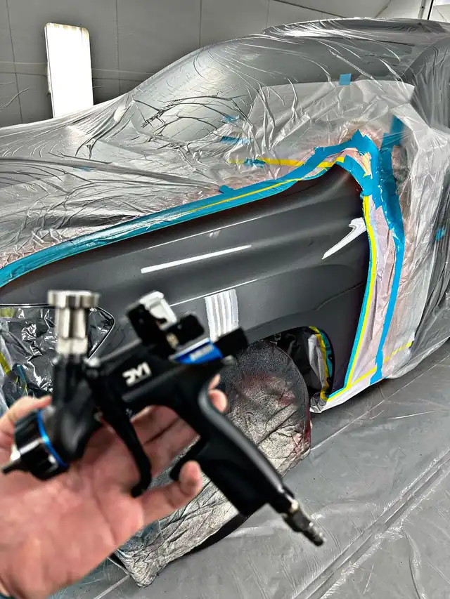 A spray gun held in front of a masked car inside the Manchester Auto Body paint booth