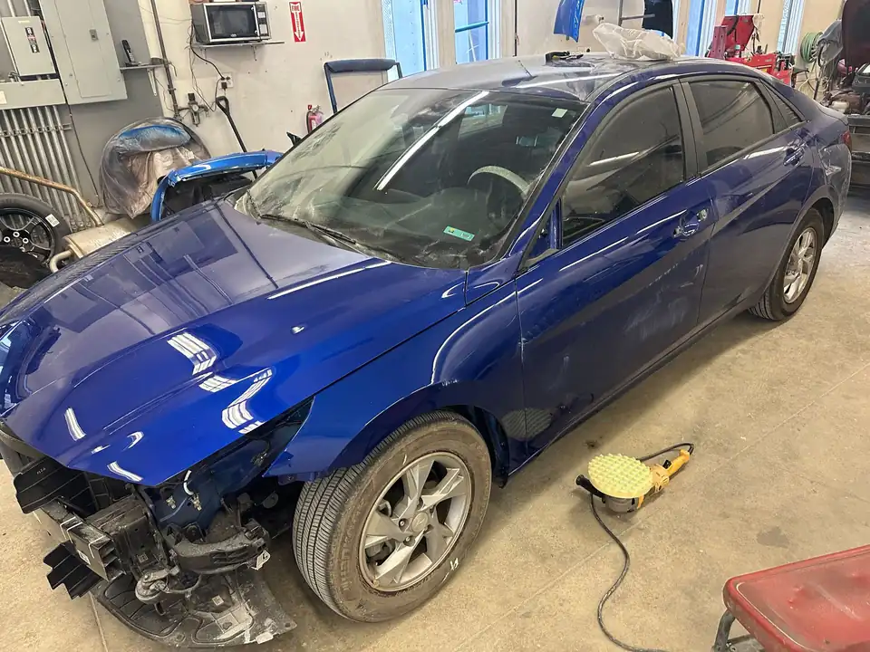 A blue Hyundai sedan mid-repair inside Manchester Auto Body, front end open and panels freshly painted
